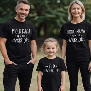 May include: Three black t-shirts with white text and a blue ribbon design. The shirts say "Proud Dada of a TID Warrior", "Proud Mama of a TID Warrior", and "TID Warrior".