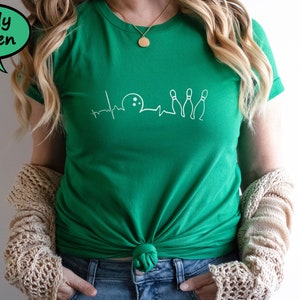 Puede incluir: Una camiseta verde kelly con un gr&aacute;fico blanco de una bola de bolos y bolos con una l&iacute;nea de latido del coraz&oacute;n. La camiseta est&aacute; anudada a la cintura.