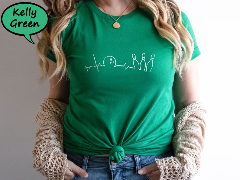 Puede incluir: Una camiseta verde kelly con un gr&aacute;fico blanco de una bola de bolos y bolos con una l&iacute;nea de latido del coraz&oacute;n. La camiseta est&aacute; anudada a la cintura.