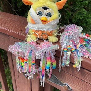 May include: A yellow and orange Furby toy with a white bandage around its head, wearing a silver chain with multiple keychains. The keychains are rainbow-coloured beaded tassels with clear plastic beads.