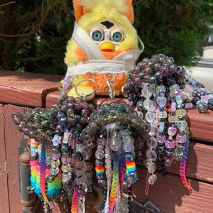 May include: Three iridescent black beaded keychain charms with rainbow sequin tails hanging from a wooden surface. A yellow and orange toy with blue eyes is in the background.