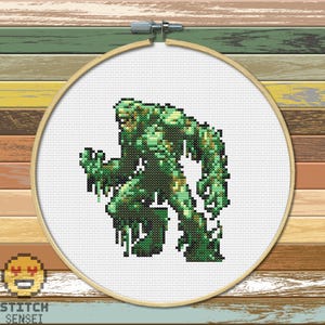 May include: Cross stitch pattern of a green monster with a large head and long arms. The monster is standing on a white background.