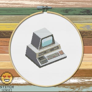 May include: Cross-stitch embroidery of a retro computer in a wooden hoop. The computer is grey and white with a screen, keyboard, and buttons. The hoop is set against a background of colourful wooden planks. The text "STITCH SENSEI" is at the bottom.