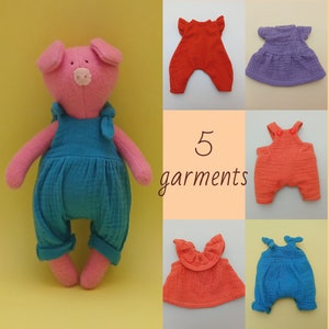 Op de afbeelding: Een roze varkenspop met een blauwe overall. De afbeelding toont ook vijf kleurrijke kledingstukken: rode en oranje overalls, een paarse jurk en een blauwe romper. De tekst "5 garments" staat in het midden.