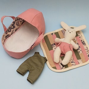 May include: A pink fabric baby carrier with a white lining, a green fabric pair of dungarees, and a white stuffed bunny wearing pink shorts lying on a patchwork quilt with pink, green, and brown fabric squares.