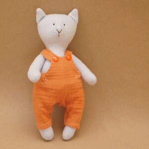 May include: A white plush cat toy wearing orange overalls with two orange buttons. The cat has black eyes and a black nose.