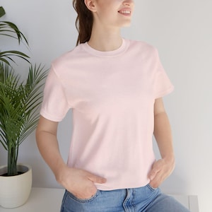 May include: A light pink crew neck t-shirt. The short-sleeved shirt is made of a soft material. The person is wearing blue jeans. A potted plant is in the background.