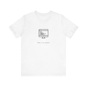 Puede incluir: Camiseta blanca con un gráfico negro de un monitor de computadora con código y la frase "Works on my machine". El diseño está centrado en la parte delantera de la camiseta.
