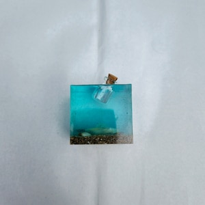 May include: A small glass bottle with a cork stopper is encased in clear blue resin, resembling a message in a bottle at the bottom of the ocean. The resin is set on a bed of brown sand.