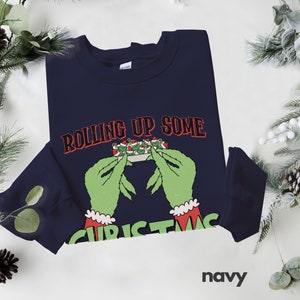 May include: Navy blue sweatshirt with a green Grinch-like hand rolling up a Christmas-themed joint. The text "Rolling Up Some Christmas" is printed on the sweatshirt.