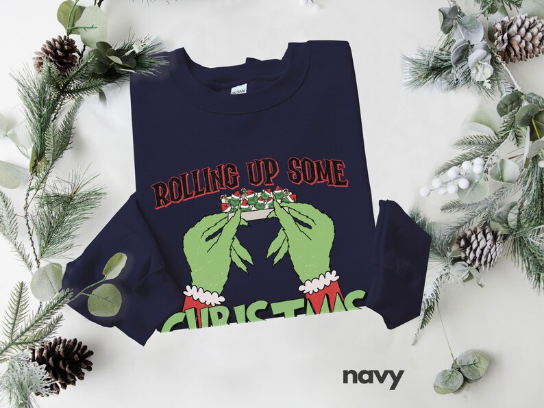 May include: Navy blue sweatshirt with a green Grinch-like hand rolling up a Christmas-themed joint. The text "Rolling Up Some Christmas" is printed on the sweatshirt.