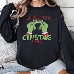 May include: A black sweatshirt with a green Grinch-like hand holding a Christmas-themed object. The text on the sweatshirt reads "Rolling Up Some Christmas Spirit".