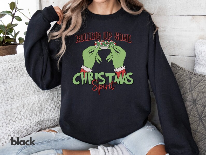 May include: A black sweatshirt with a green Grinch-like hand holding a Christmas-themed object. The text on the sweatshirt reads "Rolling Up Some Christmas Spirit".