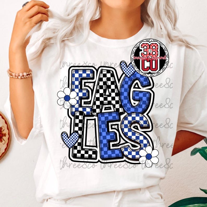 Puede incluir: Camiseta blanca con la palabra "EAGLES" en un dise&ntilde;o a cuadros azul y negro. La camiseta presenta detalles florales y de coraz&oacute;n, y un logotipo circular con el n&uacute;mero 38.