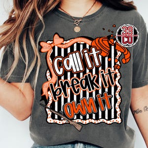 May include: Dark grey t-shirt with the text "Call it, break it, own it" in white and orange, set against a black and white striped background. The design includes a bow, shotgun, and a logo.