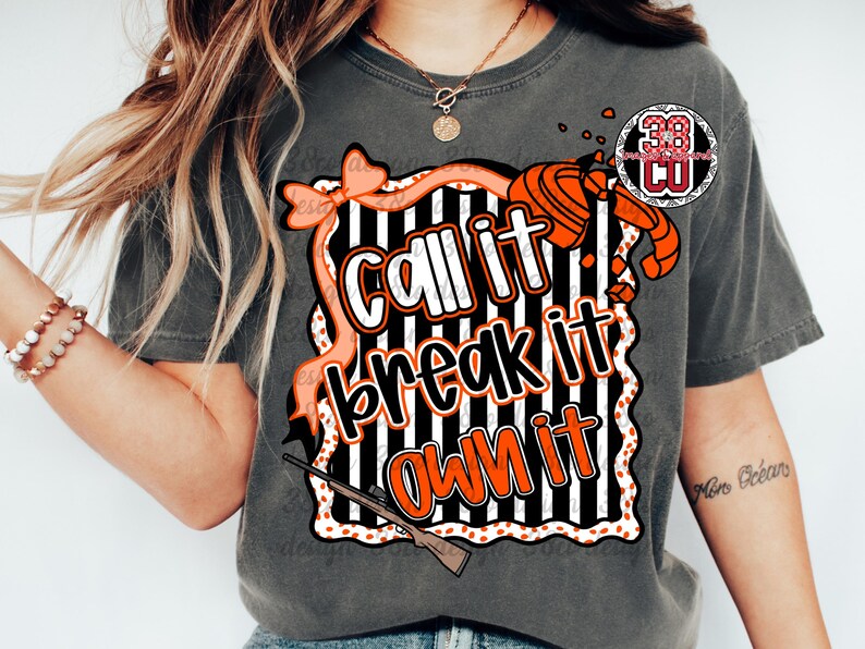 May include: Dark grey t-shirt with the text "Call it, break it, own it" in white and orange, set against a black and white striped background. The design includes a bow, shotgun, and a logo.