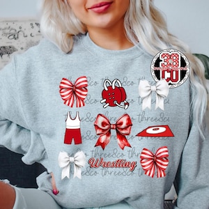 May include: A light gray sweatshirt featuring a collection of red and white bows, wrestling gear, and the word "Wrestling" in a script font. A circular logo is on the upper right. The design is a sports theme.