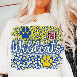 May include: An off-white t-shirt featuring a graphic design with blue and yellow leopard print patterns, a blue paw print, a yellow paw print, and the word Wildcats in blue script. A circular logo with the text 38 CO is visible on the design.