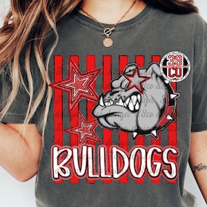 May include: A charcoal gray t-shirt featuring a graphic of a bulldog head wearing a spiked collar, two red stars, and vertical red stripes. The word BULLDOGS is printed in large white letters with a red outline at the bottom of the graphic design.