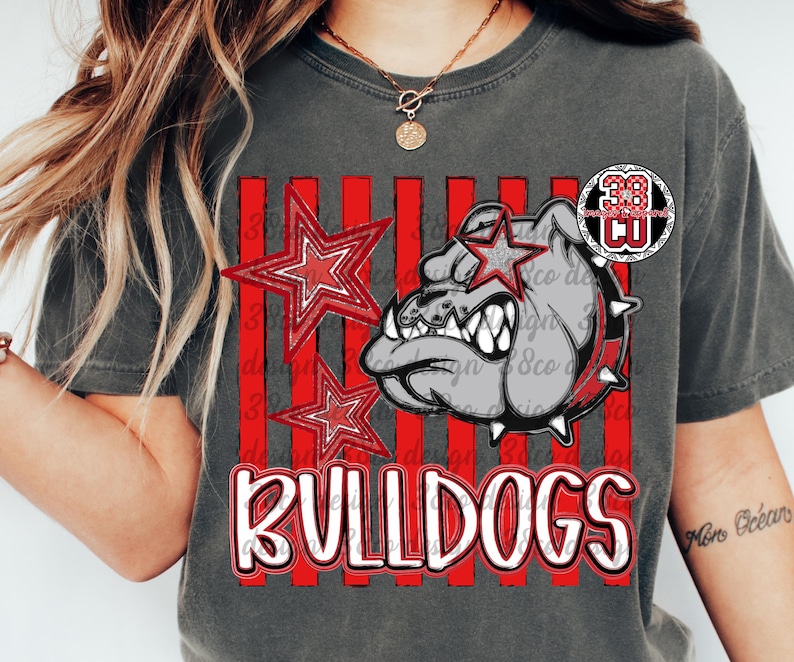 May include: A charcoal gray t-shirt featuring a graphic of a bulldog head wearing a spiked collar, two red stars, and vertical red stripes. The word BULLDOGS is printed in large white letters with a red outline at the bottom of the graphic design.