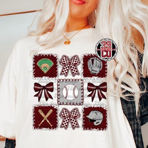 May include: Cream-colored t-shirt featuring a baseball-themed design. The design includes squares with baseball elements like a baseball field, bows, a baseball, a glove, crossed bats, and a helmet. The design is in maroon, white, and gray.