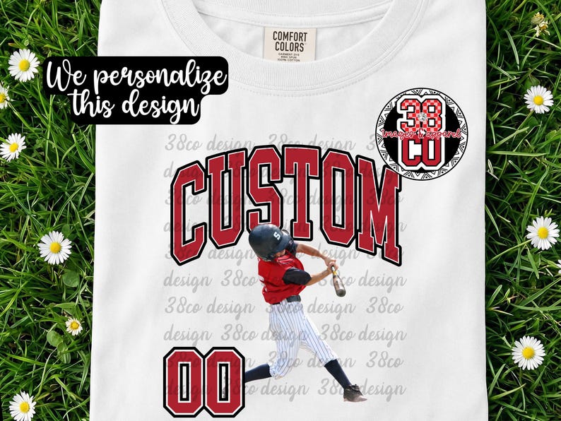 May include: White t-shirt with a baseball theme. The shirt features the word "CUSTOM" in red block letters, a baseball player graphic, and the numbers "00" in red. The text "We personalize this design" is also visible.