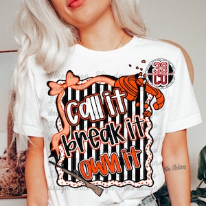 May include: White t-shirt with a graphic design featuring the text "Call it, Break it, Own it" in orange and white. The design includes a bow, a shotgun, and a target, with a black and white striped background.