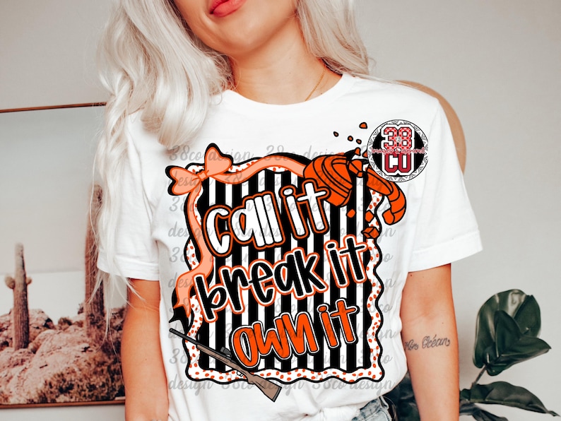 May include: White t-shirt with a graphic design featuring the text "Call it, Break it, Own it" in orange and white. The design includes a bow, a shotgun, and a target, with a black and white striped background.
