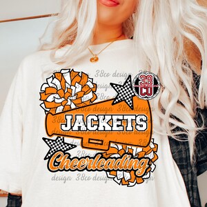Peut inclure: T-shirt blanc avec un motif de pompons orange et blanc, un mégaphone et des étoiles. Le texte sur le t-shirt indique "JACKETS" et "Cheerleading" en noir et orange. Le design comprend le texte "38co design".