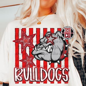 May include: A cream-colored t-shirt featuring a graphic of a grey bulldog head with a spiked collar, red and white vertical stripes, two red stars, and the word BULLDOGS in bold white letters with a red outline, plus a small circular logo.