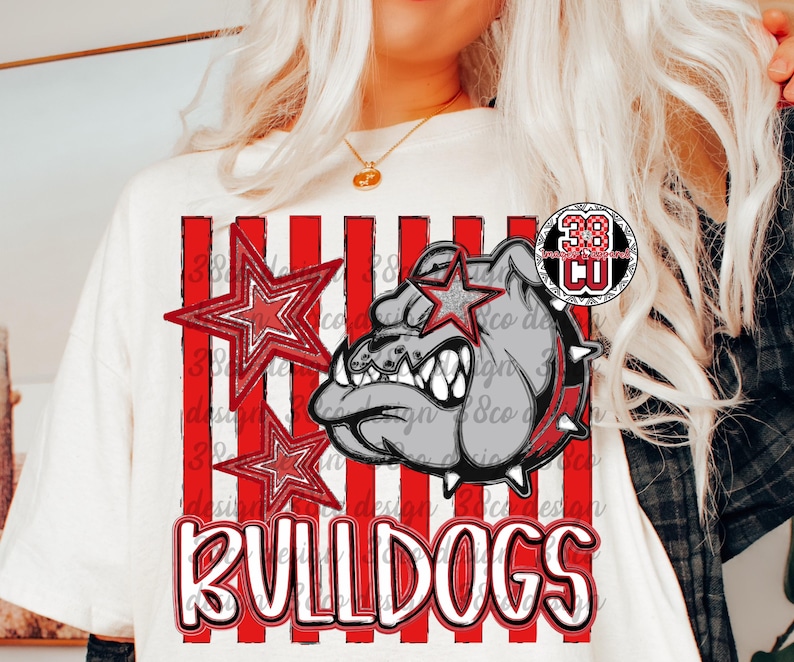 May include: A cream-colored t-shirt featuring a graphic of a grey bulldog head with a spiked collar, red and white vertical stripes, two red stars, and the word BULLDOGS in bold white letters with a red outline, plus a small circular logo.