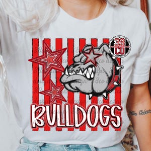 May include: A white short-sleeve t-shirt featuring a graphic of a grey bulldog wearing a spiked collar, accented by red striped vertical lines and two red stars. The text BULLDOGS is printed in large red letters at the bottom, with a 38 CO logo.