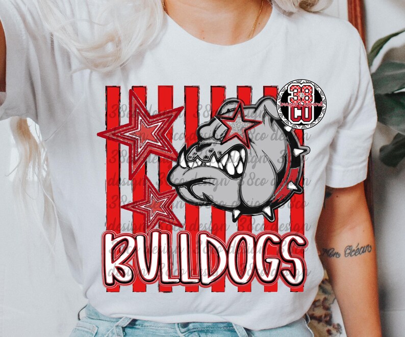 May include: A white short-sleeve t-shirt featuring a graphic of a grey bulldog wearing a spiked collar, accented by red striped vertical lines and two red stars. The text BULLDOGS is printed in large red letters at the bottom, with a 38 CO logo.