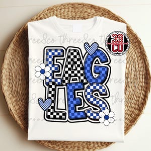 Puede incluir: Camiseta blanca con la palabra "EAGLES" en un dise&ntilde;o de letras en bloque audaz. Las letras son una mezcla de patrones de cuadros azules y negros, con detalles en azul, flores y corazones blancos. Un logotipo circular est&aacute; en la esquina superior derecha.