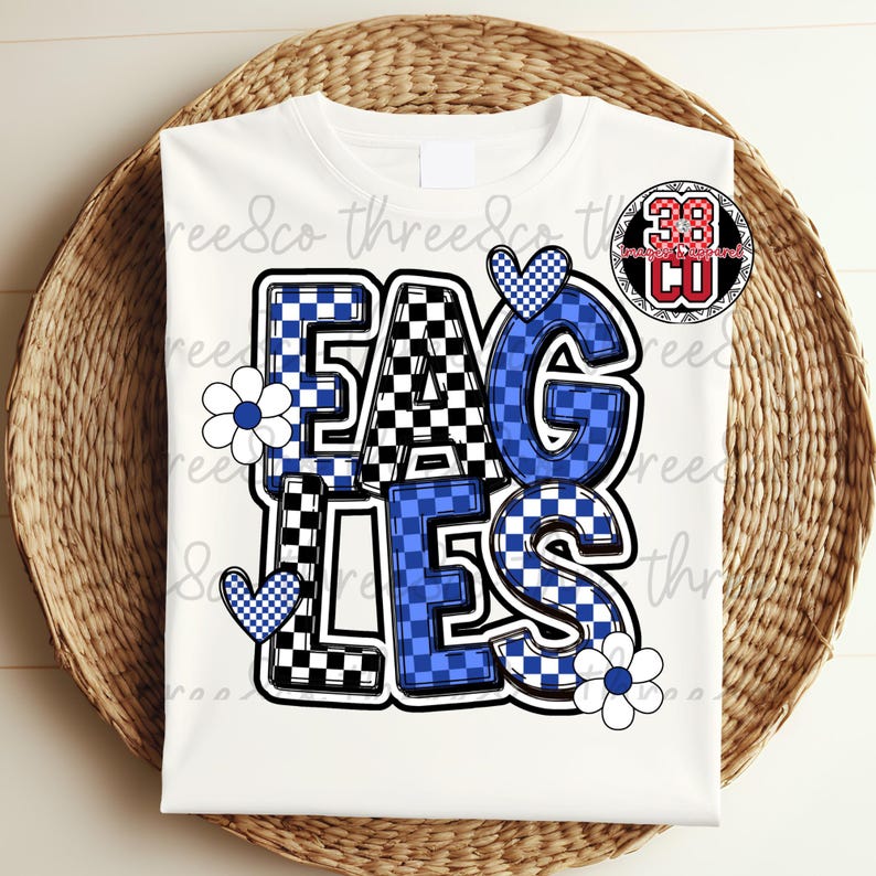 Puede incluir: Camiseta blanca con la palabra "EAGLES" en un dise&ntilde;o de letras en bloque audaz. Las letras son una mezcla de patrones de cuadros azules y negros, con detalles en azul, flores y corazones blancos. Un logotipo circular est&aacute; en la esquina superior derecha.