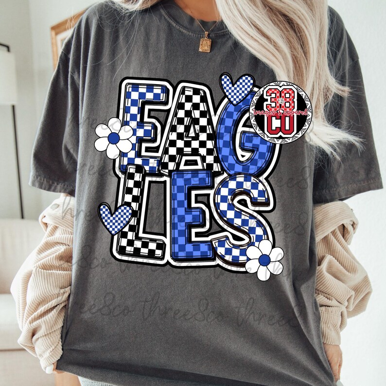 Puede incluir: Camiseta gris oscuro con la palabra "EAGLES" en un dise&ntilde;o a cuadros azul y negro. La camiseta presenta flores blancas y corazones a cuadros azules. Un logotipo circular con el n&uacute;mero "38" est&aacute; a la derecha.