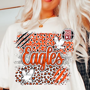 Può includere: T-shirt bianca con grafica "Eagles" arancione e bianca. Il design include uno sfondo con stampa leopardata, illustrazioni di aquile e artigli. È presente anche un logo circolare con la scritta "38 CU".