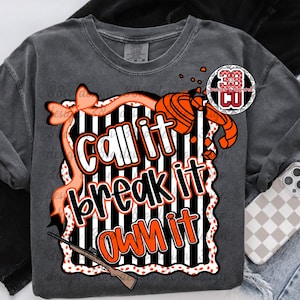 May include: Dark gray t-shirt with the text "Call it, Break it, Own it" in white and orange. The design includes a bow, a shotgun, and a target shooting graphic. The shirt is folded with a pair of jeans and a phone case.