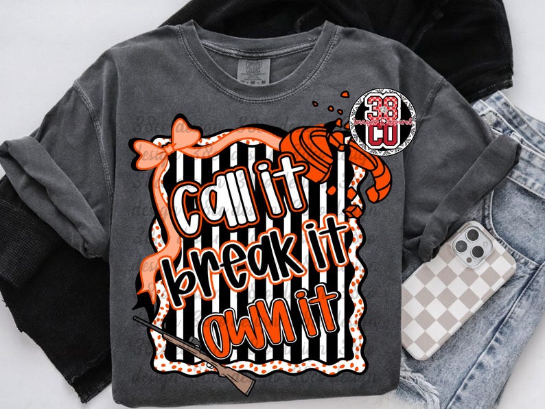 May include: Dark gray t-shirt with the text "Call it, Break it, Own it" in white and orange. The design includes a bow, a shotgun, and a target shooting graphic. The shirt is folded with a pair of jeans and a phone case.