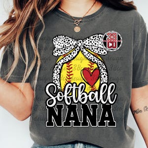 May include: Dark gray t-shirt with a softball graphic. The design features a yellow softball with a red heart, a black and white polka dot bow, and the words "Softball NANA" in white and black. A small circular graphic is above the softball.
