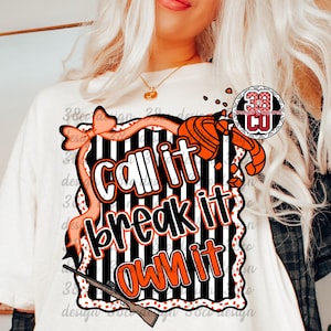 May include: Cream-colored t-shirt with a graphic that says "Call it, Break it, Own it" in orange and white. The design includes a bow, a shotgun, and a clay target. The shirt has a round neck and short sleeves.
