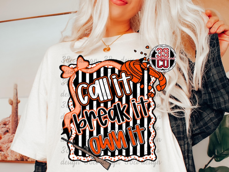May include: Cream-colored t-shirt with a graphic that says "Call it, Break it, Own it" in orange and white. The design includes a bow, a shotgun, and a clay target. The shirt has a round neck and short sleeves.