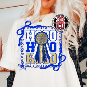 May include: Cream-colored t-shirt featuring a blue and gold Homecoming design. The design includes the word "Homecoming" on a ribbon, a star, and the letters "H-O-C-O" in a bold font. The shirt has a round neckline.