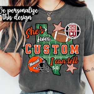 May include: A charcoal gray t-shirt featuring a green Texas-shaped graphic with a football field pattern. The design includes the text "She's from CUSTOM I can tell", an orange football helmet labeled "Mascot", a brown football, and two orange checkered stars.