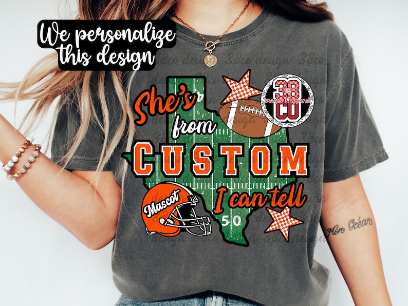 May include: A charcoal gray t-shirt featuring a green Texas-shaped graphic with a football field pattern. The design includes the text "She's from CUSTOM I can tell", an orange football helmet labeled "Mascot", a brown football, and two orange checkered stars.