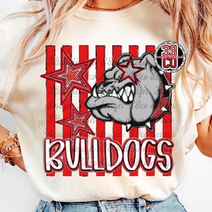 May include: A cream-colored t-shirt featuring a graphic print of a grey bulldog head with a spiked collar, two red stars, and vertical red stripes. The word BULLDOGS is printed in large white letters with a red outline at the bottom of the design.