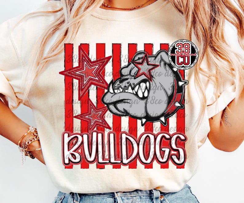 May include: A cream-colored t-shirt featuring a graphic print of a grey bulldog head with a spiked collar, two red stars, and vertical red stripes. The word BULLDOGS is printed in large white letters with a red outline at the bottom of the design.