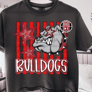 May include: A charcoal gray short-sleeve t-shirt featuring a graphic print of a bulldog head with a star over one eye, two red stars, and vertical red stripes. The text "BULLDOGS" is printed in white with a red outline at the bottom of the design.