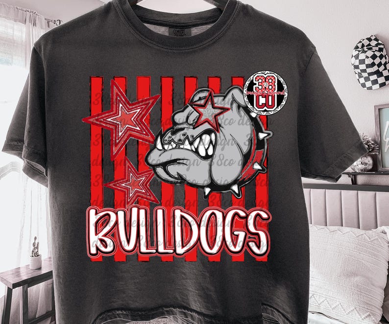 May include: A charcoal gray short-sleeve t-shirt featuring a graphic print of a bulldog head with a star over one eye, two red stars, and vertical red stripes. The text "BULLDOGS" is printed in white with a red outline at the bottom of the design.