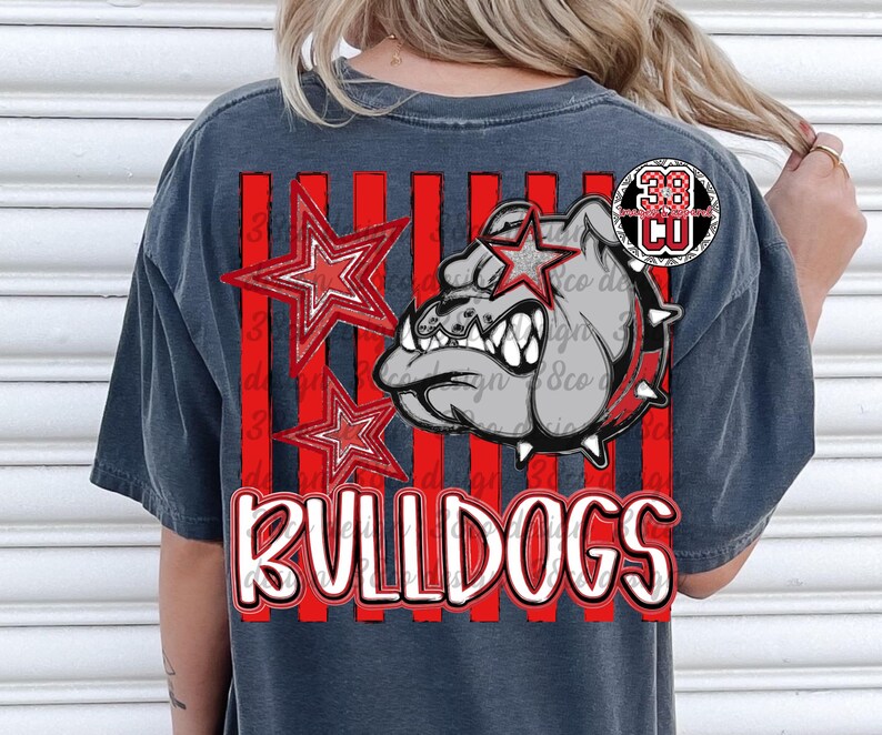 May include: A charcoal gray t-shirt featuring a graphic print of a bulldog head with a spiked collar, two red stars, and vertical red stripes. The word BULLDOGS is printed in large white letters with a red outline at the bottom of the design.